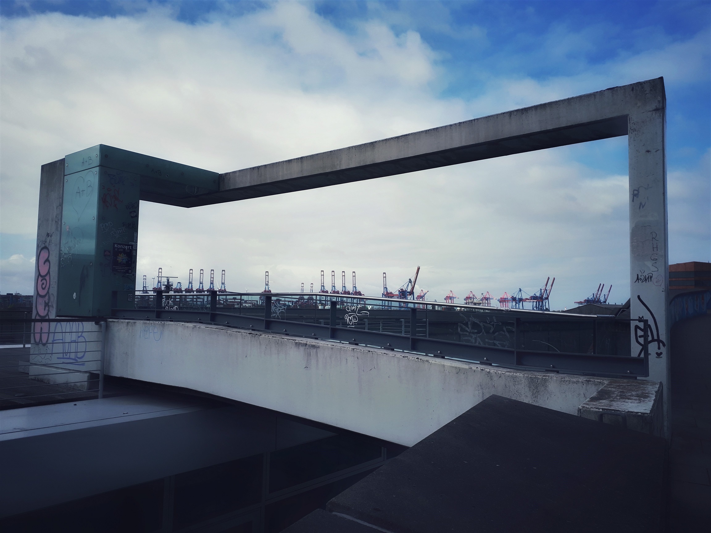 Durch einen Betonrahmen fotografiert. Im Hintergrund sind die Kräne an der gegenüberliegenden Elbseite zu sehen. Sie scheinen vom Beton eingerahmt und erinnern ein wenig an erhobene Taktstöcke.